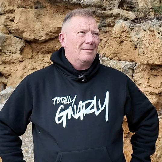 Man wearing a black hoodie with 'Totally Gnarly' text against a stone wall.