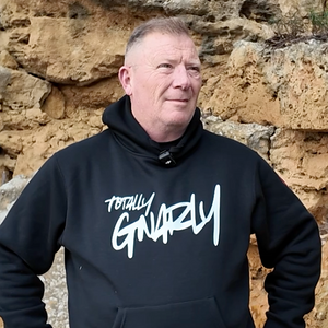 Man wearing a black hoodie with 'Totally Gnarly' text against a stone wall.