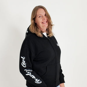 Person wearing a black hoodie with text, standing against a white wall with plants on either side.