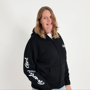 Person wearing a black hoodie with text, standing against a white background with plants on either side.