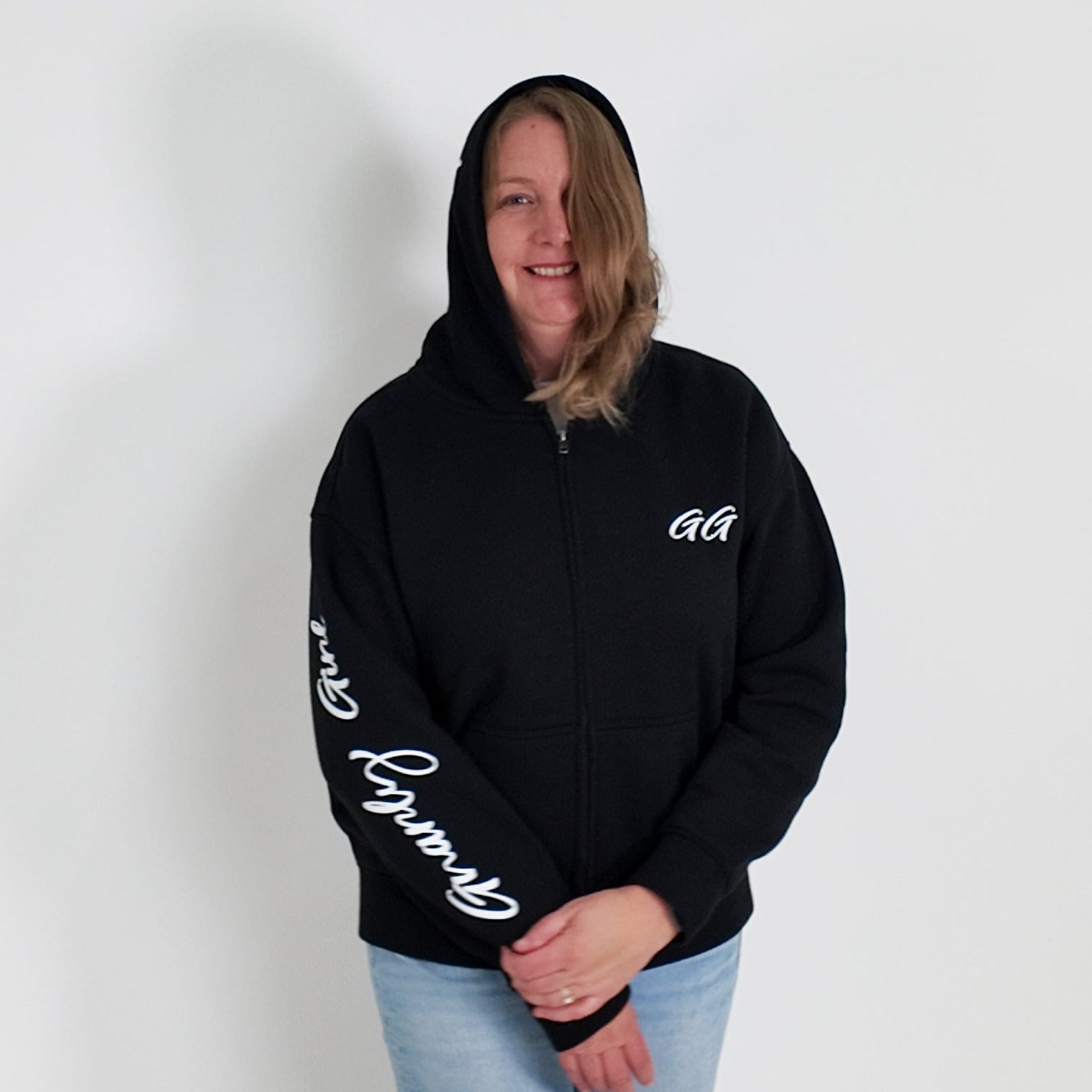 Person wearing a black hoodie with white text, standing against a plain white wall with a plant on the left.